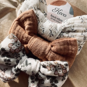 以下が含まれることがあります： 花柄の生地でできたヘアボウが3つ、中央に茶色のボウがあります。ボウは三角形に配置されています。ラベルには「Keva Noeuds enfants fait avec amour」と書いてあります。