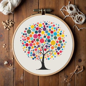 May include: A cross-stitch embroidery of a tree with a black trunk and branches, and a colorful canopy of various sized circles in shades of red, orange, yellow, green, blue, and purple. The embroidery is set in a wooden hoop.