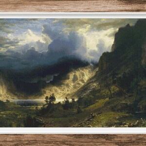 May include: A framed landscape painting depicts a mountain valley scene. The artwork features a dramatic sky with dark clouds and sunlight illuminating the valley. The painting is set in a white frame, with a wooden background.