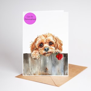May include: A greeting card featuring a watercolor illustration of a Cavalier King Charles Spaniel dog peeking over a wooden fence, holding a red rose. The card has the text "Can be Personalised" in a pink circle.