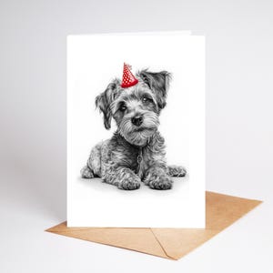 May include: A black and white illustration of a small dog wearing a red party hat with white polka dots. The dog is lying down on a white card, which is resting on a brown envelope. The image is a greeting card.