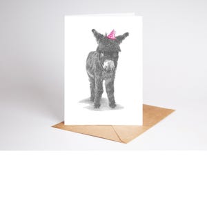 May include: A greeting card with a black and white drawing of a donkey wearing a pink party hat with white spots. The card is on a white background, placed on a brown envelope.