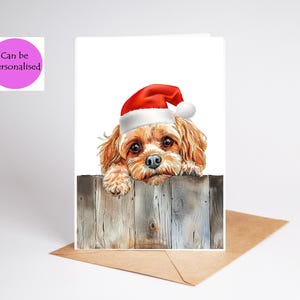 Peut inclure: Une carte de vœux avec une illustration à l'aquarelle d'un chien marron portant un bonnet de Père Noël rouge et blanc, posant ses pattes sur une clôture en bois gris. La carte inclut un badge rose avec le texte Can be Personalised.