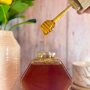 May include: A clear glass jar with a hexagonal shape filled with honey. A wooden honey dipper with a drip of honey is shown above the jar.