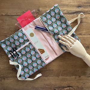 Peut inclure: Trousse à stylos et crayons à motifs floraux bleus et roses, avec un intérieur rose. Elle contient des stylos, des crayons et une boîte de crayons "plar". Une paire de lunettes roses et une main en bois sont également visibles. Fermeture éclair.