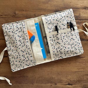 Pode incluir: Capa de livro aberta com um padrão floral em cinza, azul marinho e laranja. A capa tem um bolso com zíper, porta-canetas e uma fita. Dentro estão livros e canetas. A capa está sobre uma superfície de madeira.