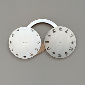 May include: Two silver watch dials with black numerals and markings, one indicating hours and the other minutes. The dials are connected by a light brown wooden piece and a silver ring.