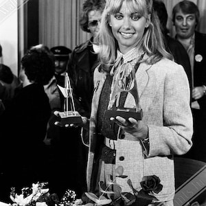 May include: A woman with long blonde hair holds two silver awards in front of a crowd of people. She is wearing a light brown blazer over a black sweater and a white shirt. She is smiling.