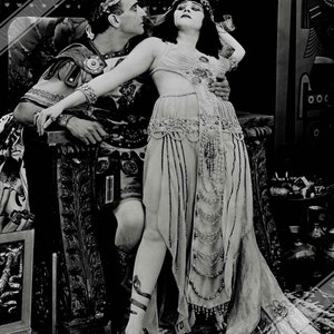 Pode incluir: Uma fotografia a preto e branco de um casal numa pose dramática. A mulher está vestida com um vestido de contas e um adereço de cabeça, enquanto o homem está vestido com uma túnica e uma coroa de louro. Eles estão sentados num trono com talhas ornamentadas.