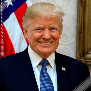 May include: A portrait of a man in a dark blue suit with a white shirt and a blue patterned tie. He is smiling and wearing a small American flag pin on his suit jacket. The American flag is visible in the background.