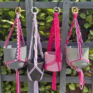 May include: Four macrame plant hangers with different coloured pots. The hangers are made with white and pink yarn and have wooden rings at the top. The pots are grey, pink, black, and grey with white speckles.