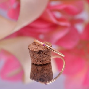 Puede incluir: Un anillo dorado con una pequeña gema rosa y redonda. El anillo está colocado sobre un corcho, con un fondo borroso rosa y blanco. El anillo se refleja en una superficie espejada.