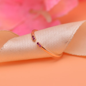 May include: A delicate gold ring with two small pink gemstones. The ring is open and sits on a white fabric background.