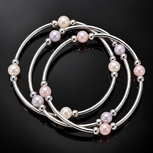 May include: A set of three interlocking bangle bracelets made of polished silver-tone metal. Each bracelet features a series of smooth, round beads in shades of white, soft pink, and pale lavender, interspersed with small, shiny silver-tone spacer beads.
