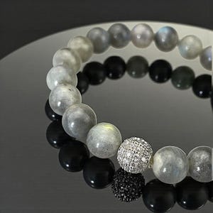 May include: A beaded bracelet featuring a mix of gray, black, and silver beads. The bracelet includes a silver bead encrusted with small, sparkling stones. The beads vary in size and texture, creating a unique and elegant accessory.