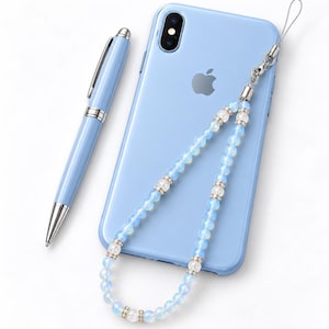 May include: A light blue smartphone with a dual-lens camera and an Apple logo, accompanied by a matching light blue ballpoint pen with silver accents and a beaded phone charm featuring translucent opalescent beads and silver-toned spacers.