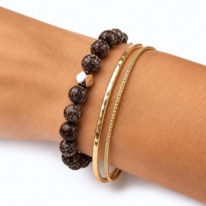 May include: A set of three bracelets consisting of one beaded bracelet with dark brown speckled beads and a single silver-toned geometric bead, alongside two thin gold-toned metal bangles, one with a hammered texture and one with a beaded edge.