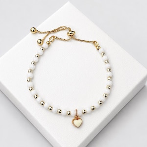 May include: A gold-toned adjustable bracelet featuring a series of alternating white spherical beads and small gold-toned metal beads. A single heart-shaped charm with a cream-coloured centre and a gold-toned border hangs from the centre of the bracelet.