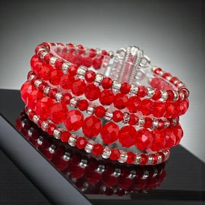 May include: A red beaded bracelet with a silver clasp. The bracelet features multiple strands of faceted red beads interspersed with smaller silver beads. The bracelet is displayed on a reflective surface.