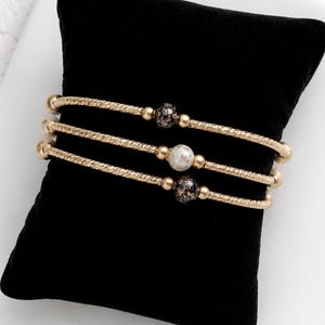 May include: Three gold-toned bracelets featuring a twisted metal texture. Each bracelet is accented with a single central bead: the top and bottom beads are black with gold flecks, while the middle bead is white with brown marble-like veining.