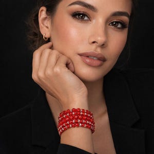 May include: A red beaded bracelet with multiple strands and small, clear accent beads. The bracelet is worn on a wrist, with a black background. The bracelet appears to be made of small, round beads.
