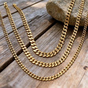Könnte beinhalten: Drei goldfarbene Kettenhalsketten mit unterschiedlich großen Gliedern werden auf einer verwitterten Holzoberfläche präsentiert. Die Ketten sind in einer geschichteten Anordnung angeordnet, mit einem Naturstein im Hintergrund. Die Halsketten sind ein warmes, glänzendes Gold.