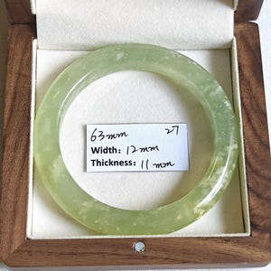 Könnte beinhalten: Ein blassgrüner, durchscheinender Armreif mit weißen, gesprenkelten Einschlüssen. Ein weißes Papieretikett im Inneren des Armreifs zeigt den Text 63mm, 27, Width: 12mm und Thickness: 11mm. Der Armreif liegt in einer quadratischen Holzschatulle.