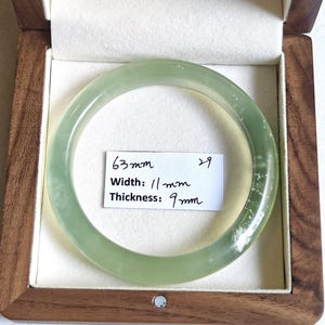Könnte beinhalten: Ein glatter, durchscheinender hellgrüner Armreif mit feinen weißen Einschlüssen. Ein weißes Papieretikett im Inneren des Kreises zeigt den Text 63mm, Width: 11mm und Thickness: 9mm, was die genauen Maße des Schmuckstücks angibt.
