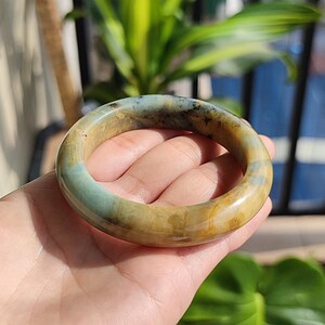May include: A jade bangle bracelet in shades of green, tan, and brown. The polished bracelet has a smooth surface and a natural stone appearance. The bangle is held in a hand. The bracelet's diameter is approximately 6.5 cm.