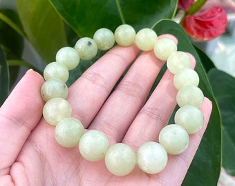 May include: A light green beaded bracelet. The bracelet is made of round, polished beads. The beads have a soft, matte finish and a pale green color. The bracelet is held in a hand against a background of green leaves and a red flower.