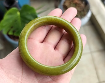 May include: A jade bangle bracelet in a light olive green hue. The smooth, circular bracelet is held in a hand, showcasing its polished surface. The bangle's color is consistent throughout, with subtle variations in tone. A piece of jewelry.