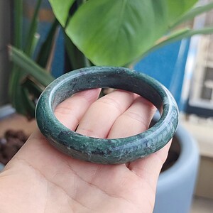以下が含まれることがあります： 濃い緑色の翡翠バングルブレスレット。ブレスレットは、より濃い緑色と黒色の縞模様を持つ、滑らかで研磨された表面を持っています。円形のブレスレットは、クラシックなジュエリーです。