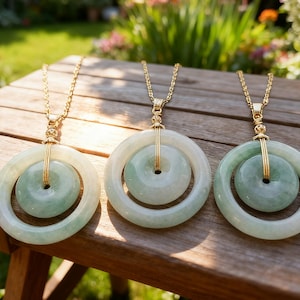 May include: Three jade pendant necklaces with gold chains rest on a wooden surface. Each pendant features a light green, circular jade stone within a larger, matching circular frame. The necklaces are displayed outdoors.