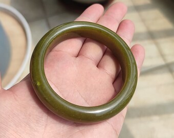 May include: Olive green jade bangle bracelet. The smooth, polished surface reflects light, highlighting the natural variations in the stone. The circular design is a classic jewelry style. This bracelet is a timeless accessory.