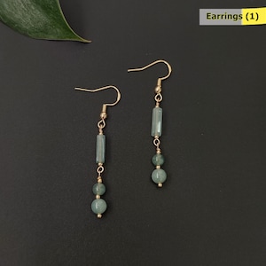 May include: A pair of dangle earrings with gold-colored hooks. Each earring features a light blue rectangular bead, two round beads, and gold-colored connecting elements. The earrings are displayed on a dark background. The text "Earrings (1)" is in the upper right corner.