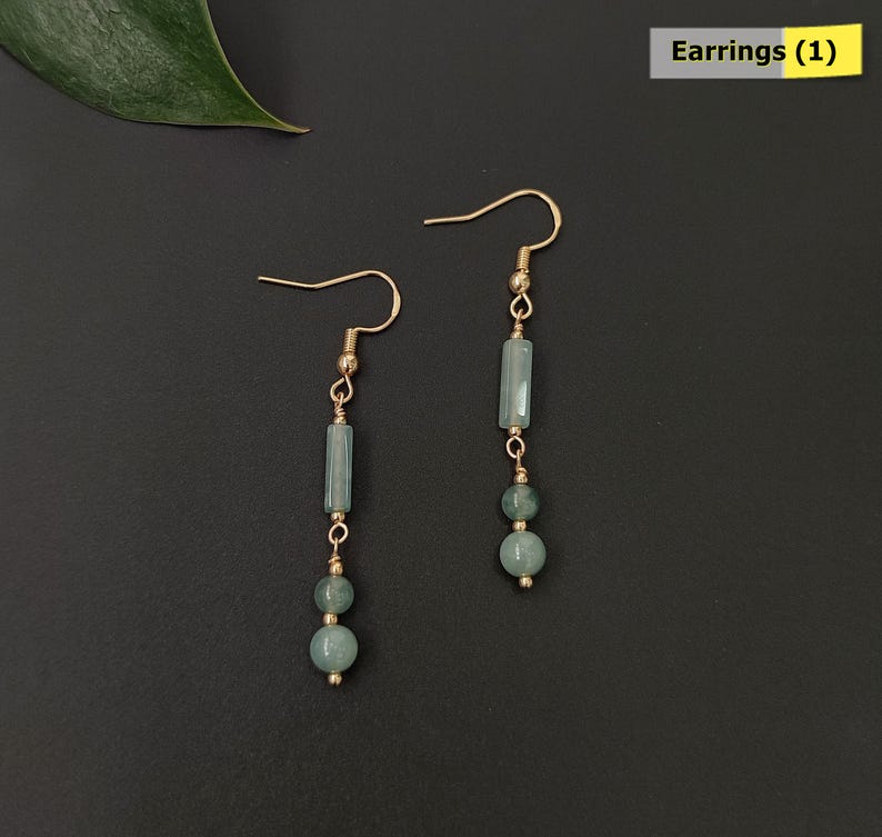 May include: A pair of dangle earrings with gold-colored hooks. Each earring features a light blue rectangular bead, two round beads, and gold-colored connecting elements. The earrings are displayed on a dark background. The text "Earrings (1)" is in the upper right corner.