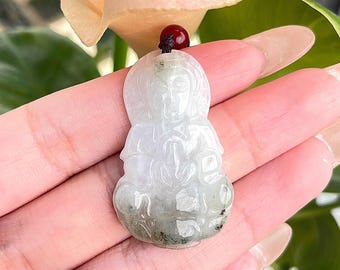 May include: A white jade pendant carved with a detailed depiction of a seated figure. The pendant has a green and white mottled colour and is attached to a dark brown cord.