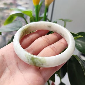 以下が含まれることがあります： 緑色のマーブル模様が入った白い翡翠のバングルブレスレット。 ブレスレットの滑らかで磨かれた表面は、緑の葉と花の背景に対して表示されます。 このエレガントなジュエリーは、クラシックなアクセサリーです。