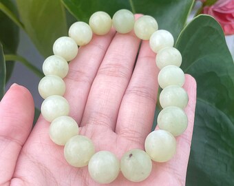 May include: A light green beaded bracelet with large, round beads. The bracelet is held in a hand, with a background of green leaves and a pink flower. The beads have a smooth, polished appearance.