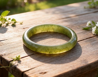 May include: A round, olive-green jade bracelet rests on a weathered wooden surface. The polished stone reflects light, showcasing its translucent quality. The bracelet's smooth surface and natural color make it a classic piece of jewelry. The bracelet is approximately 2.5 inches in diameter.
