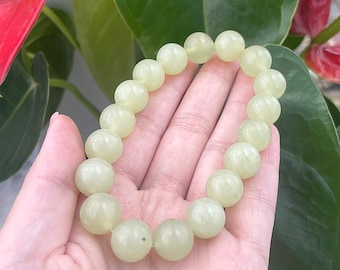 May include: A light green jade bead bracelet. The bracelet is composed of large, round, polished beads. The beads are a pale, translucent green color. The bracelet is held in a hand, with a background of green leaves and a red flower.
