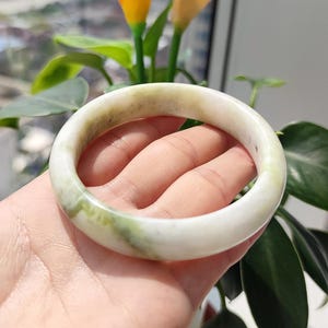 以下が含まれることがあります： 滑らかで磨かれた表面を持つ翡翠のバングルブレスレット。ブレスレットは白と緑の色合いが混ざり合い、微妙な色の変化があります。円形のデザインはシンプルでエレガントで、日常使いに適しています。