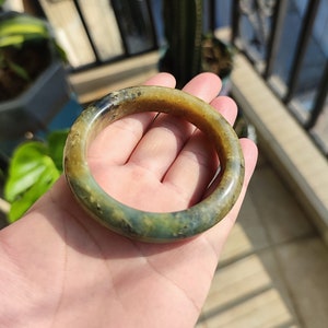 Peut inclure: Un bracelet jonc en jade aux teintes vertes, marron et noires. La surface polie du bracelet reflète la lumière, mettant en valeur les variations naturelles de couleur. Le bracelet est de forme circulaire.