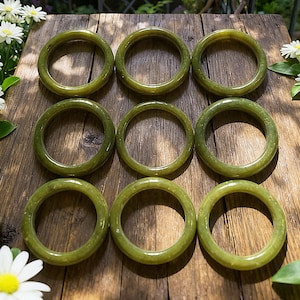 May include: Nine olive green jade bangles displayed on a wooden surface. The circular bangles have a smooth, polished finish. White flowers and greenery are visible in the background, enhancing the natural aesthetic.