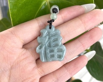Puede incluir: Un collar con un colgante de jade verde tallado con caracteres chinos. El colgante está en un cordón negro con una cuenta de jade verde.