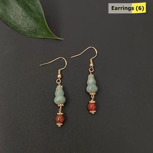 May include: A pair of gold-colored dangle earrings. Each earring features a light green gourd-shaped bead, a red-brown bead, and gold-colored accents. The earrings are displayed on a black surface. The text "Earrings (6)" is in the top right corner.