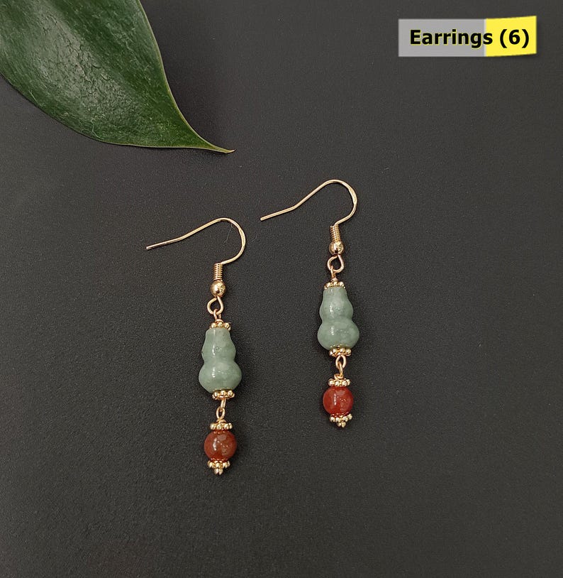 May include: A pair of gold-colored dangle earrings. Each earring features a light green gourd-shaped bead, a red-brown bead, and gold-colored accents. The earrings are displayed on a black surface. The text "Earrings (6)" is in the top right corner.
