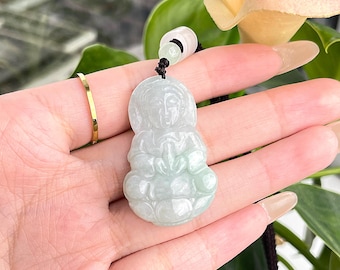 May include: A jade green pendant featuring a carved figurine of a person sitting with their hands clasped in front of them. The pendant is on a black cord.