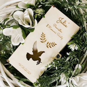 Puede incluir: Una caja de madera con un recorte de paloma y el texto "Julia - Pierwsza Komunia Swieta 19.06.2021" en letras doradas. La caja est&aacute; rodeada de flores blancas y hojas verdes.