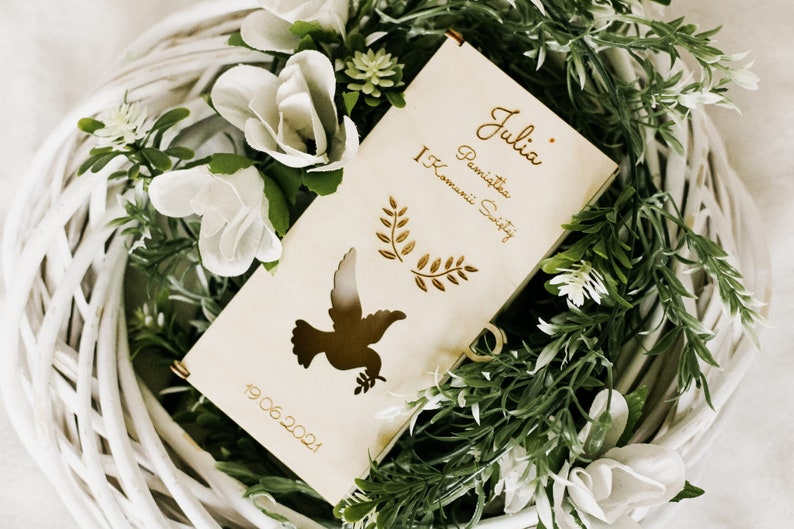 Puede incluir: Una caja de madera con un recorte de paloma y el texto "Julia - Pierwsza Komunia Swieta 19.06.2021" en letras doradas. La caja est&aacute; rodeada de flores blancas y hojas verdes.